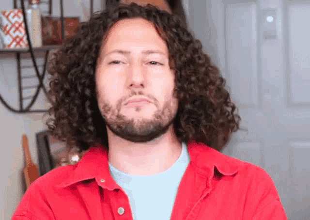 a man with curly hair and a beard wearing a red shirt is making a funny face .