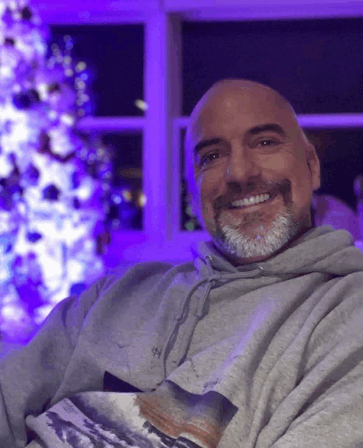 a man wearing a grey hoodie is smiling in front of a christmas tree