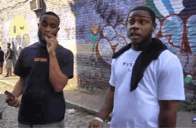 two men are standing in front of a brick wall with graffiti on it and one is wearing a shirt that says just trap
