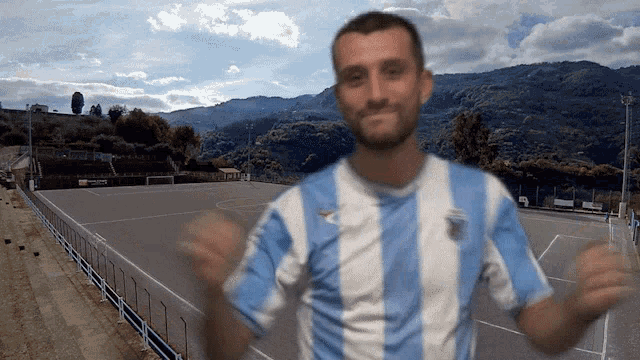 a man in a blue and white striped shirt is standing in front of a soccer field with mountains in the background