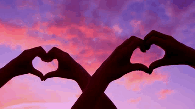 two hands are making a heart shape in front of a sunset sky .