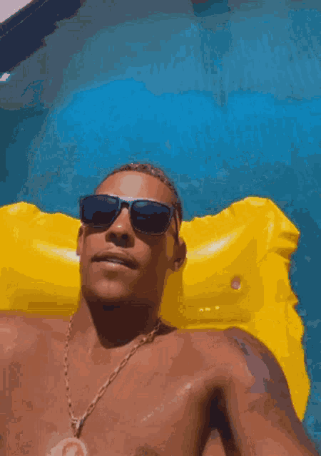 a shirtless man wearing sunglasses and a gold chain is laying on a yellow raft in a pool .
