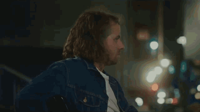 a man with long hair and a beard is wearing a blue denim jacket and white shirt .