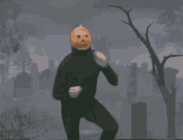 a man in a black suit with a pumpkin on his head is standing in a cemetery .