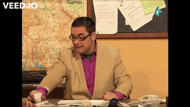 a man in a suit sits at a desk in front of a map that says veed.io on the bottom