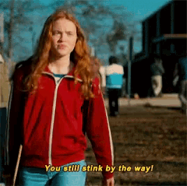 a woman in a red jacket is walking down a street and says you still stink by the way !