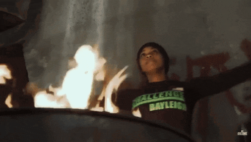 a person wearing a challenge bayleigh shirt is standing in front of a fire .