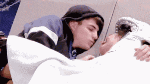 a man in a hoodie is kissing another man on the cheek while laying on a bed .