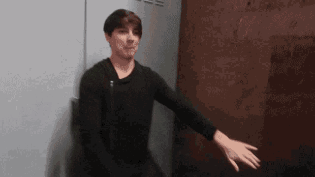 a young man in a black shirt is standing in front of a wall with his hand outstretched .