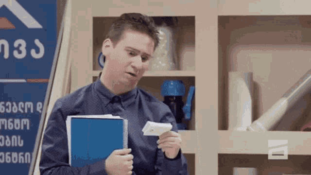 a man in a blue shirt is holding a notebook and a piece of paper in his hand