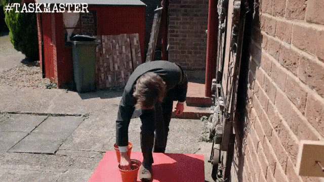 a man is pouring something into a bucket in front of a brick wall with # taskmaster written on the bottom