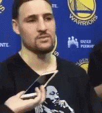 a man with a beard is holding a cell phone in front of a golden state warriors sign .