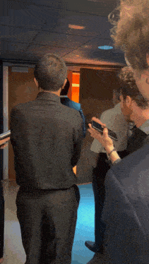 a man in a black suit holds a cell phone in his right hand