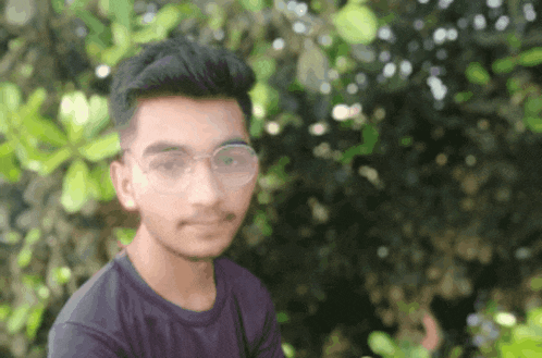 a young man wearing glasses and a purple shirt is standing in front of a tree .