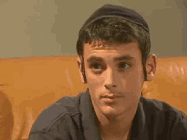 a young man wearing a hat is sitting on an orange couch looking at the camera .
