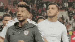 two men are standing next to each other on a soccer field . one of the men is wearing a white shirt .