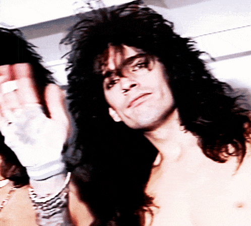 a shirtless man with long black hair looks at the camera with his hand wrapped in bandages