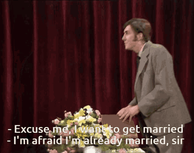 a man in a suit stands in front of a flower arrangement and says " excuse me "