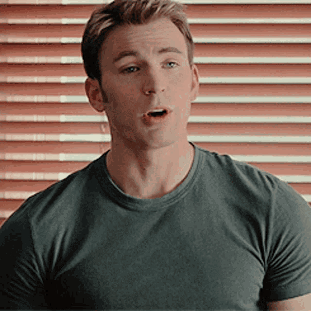 a man wearing a grey shirt is standing in front of a window with blinds .