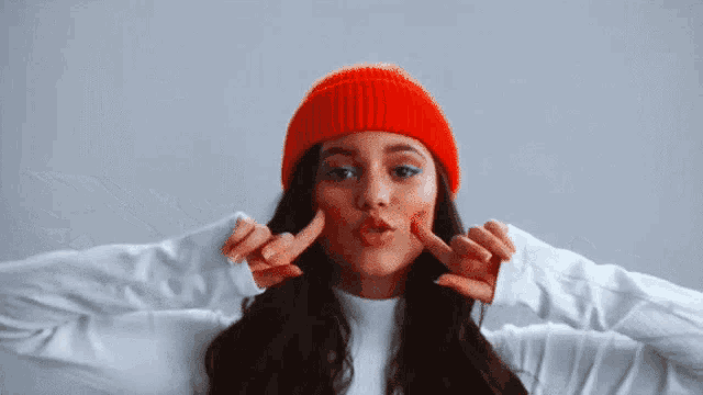 a woman wearing a red beanie and a white sweater is making a face with her hands .