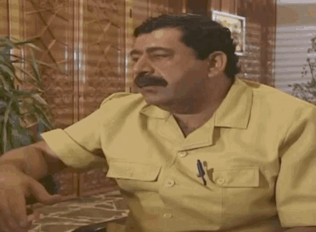 a man in a yellow shirt with a mustache is sitting at a table .