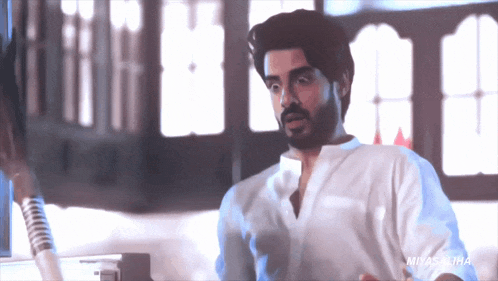 a man with a beard wearing a white shirt is standing in front of a window in a room