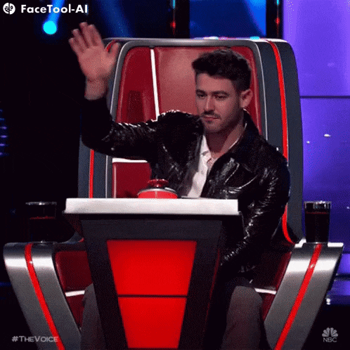 a man in a leather jacket is waving while sitting in a chair on nbc 's the voice