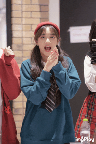 a girl wearing a blue sweatshirt and a red hat applauds in front of a bottle of water