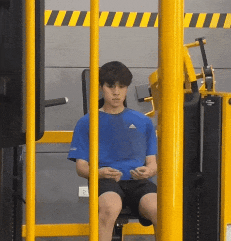 a man in a blue adidas shirt is sitting on a machine in a gym