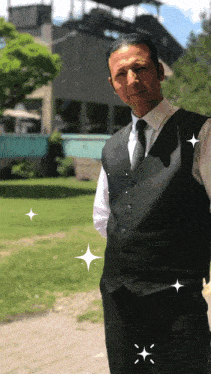 a man in a black vest and tie is standing in front of a building