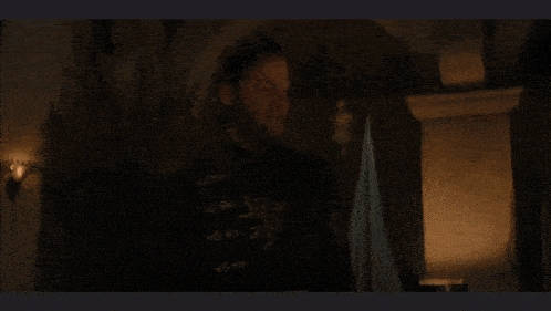 a man with a beard is standing in a dark room in front of a fireplace