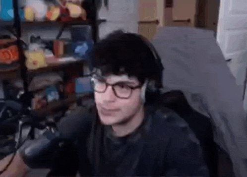 a young man wearing glasses and headphones is sitting in front of a microphone in a bedroom .