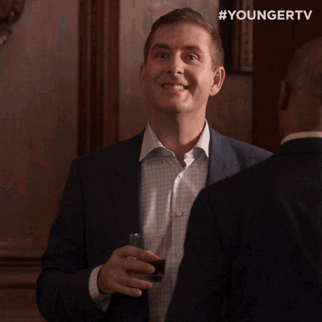 a man in a suit smiles while holding a glass in front of a younger tv logo
