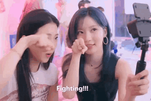 a couple of girls are standing next to each other in front of a camera and pointing at the camera .