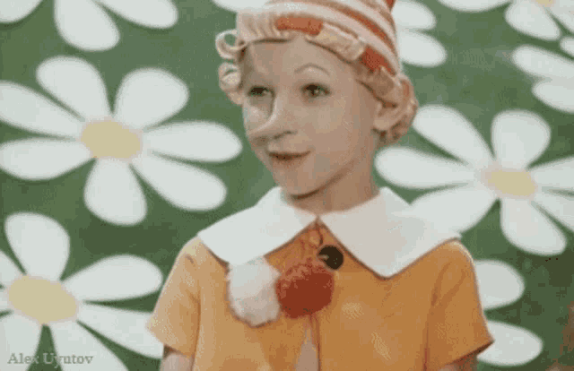 a little girl in a yellow dress and a red hat is standing in front of a wall of daisies .
