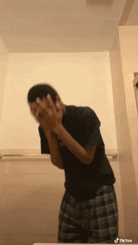 a man in a black shirt and plaid shorts is covering his face with his hands in a bathroom