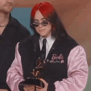 billie eilish is wearing a barbie jacket and sunglasses while holding a trophy .