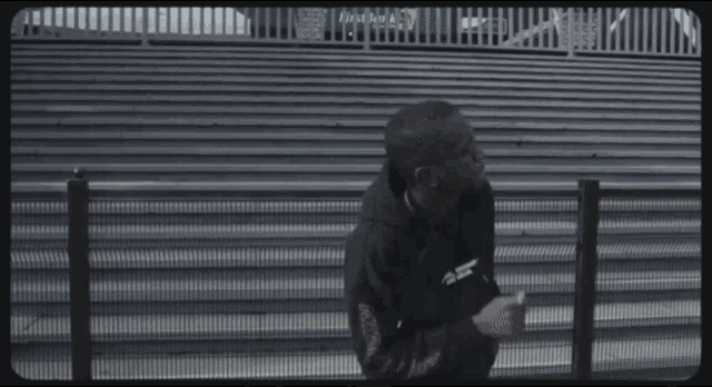 a man in a black hoodie stands in front of a fence that says first bank