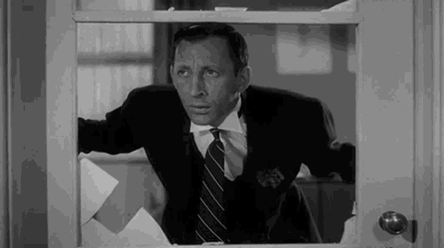 a black and white photo of a man in a suit and tie looking out of a window .