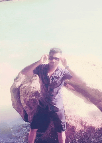 a man wearing sunglasses is standing next to a large rock in the water .