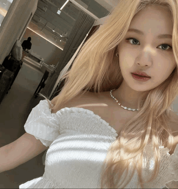 a blonde woman wearing a white off the shoulder top and a pearl necklace