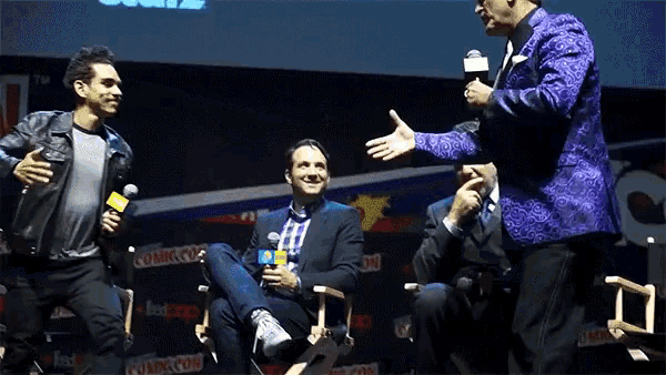 a man in a purple suit is talking into a microphone at a comic con event