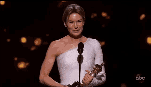 a woman in a white dress is holding an oscar statue and speaking into a microphone .