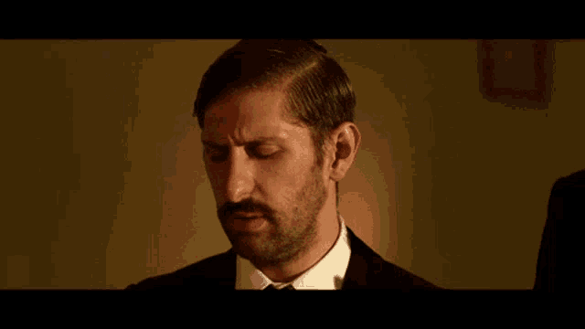 a man with a beard wearing a suit and tie is looking down