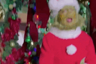 a person dressed as grinch is standing in front of a christmas tree and making a funny face .