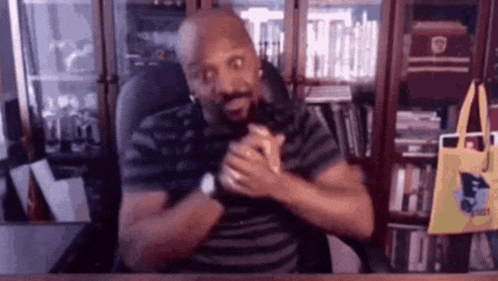 a man is sitting at a desk in front of a bookshelf and making a funny face .