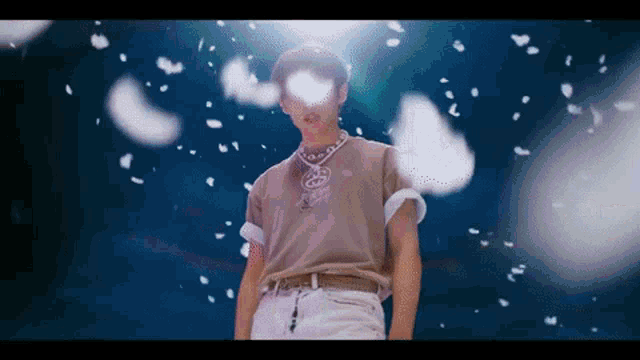 a man in a brown shirt and white pants is standing in a dark room with feathers falling around him .