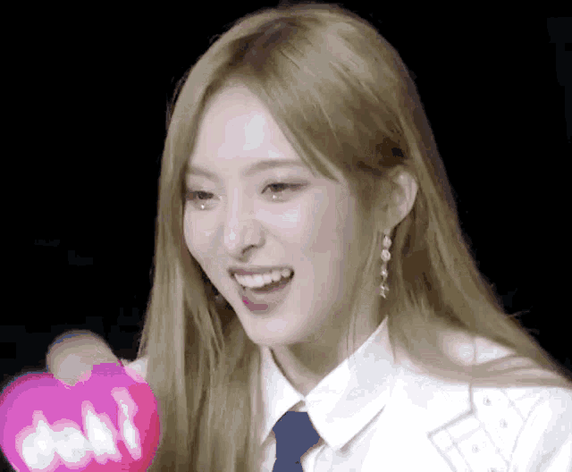 a woman with long blonde hair is smiling and holding a pink heart in her hands .