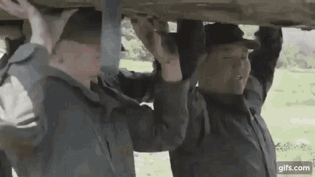 two men are standing under a car and holding onto it .