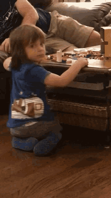 a young boy in a blue shirt is kneeling on the floor in front of a table with toys on it